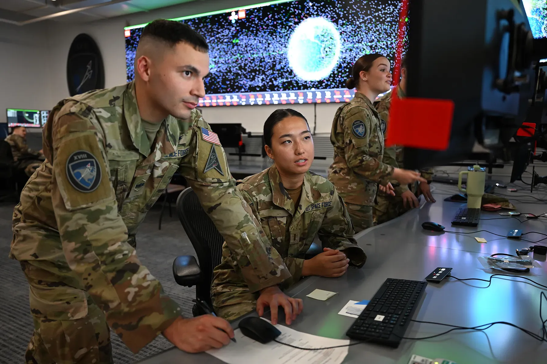 Two U.S. Space Force members collaborate at a computer console in an operations center, with a large background screen displaying a space monitoring graphic.